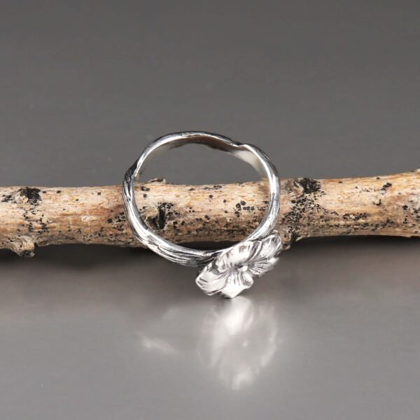 Close-up of a handcrafted sterling silver flower ring with a detailed floral design on a textured band, placed against a smooth gray background.